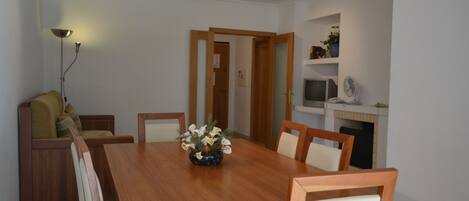 A dining area with a wooden table, chairs, and a vase of flowers. There is a sofa with cushions and a lamp on the left. A television and a shelf with decorative items are on the right wall.