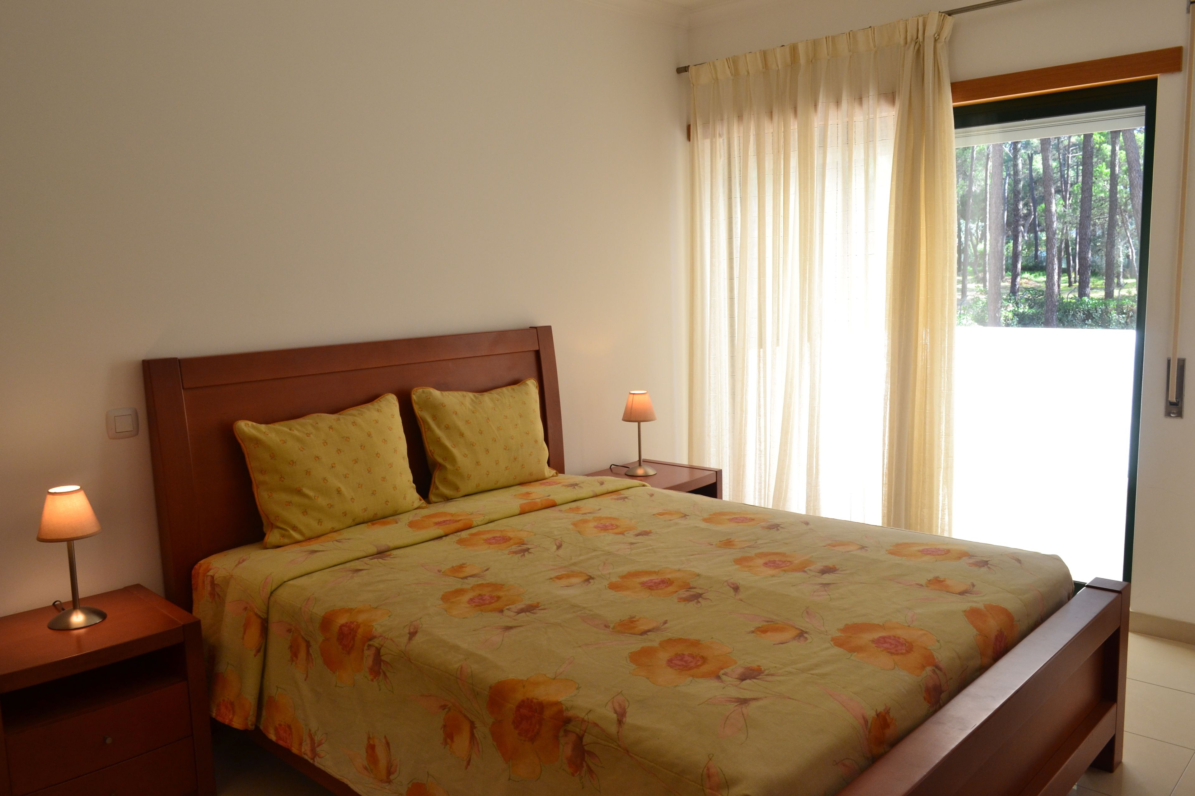 A bedroom with a wooden bed, two nightstands with lamps, a window with curtains, and a view of trees outside.