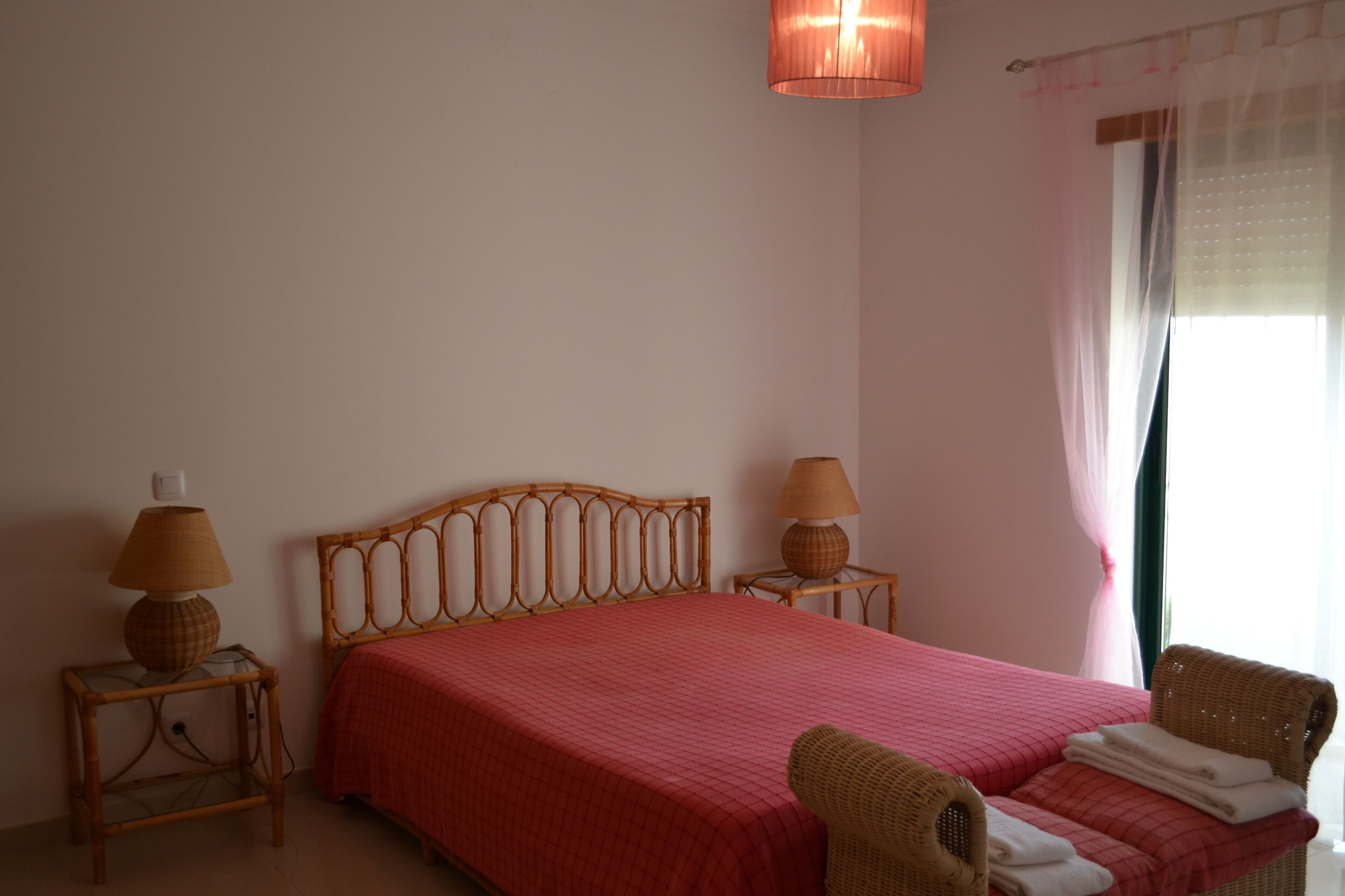 A bedroom with a wooden bed, two bedside tables with lamps, a wicker chair, and a window with curtains.