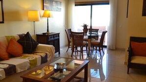A living room with a sofa, a glass coffee table, a dining table with chairs, and a lamp. - Eden Village - Apartments (Loulé)