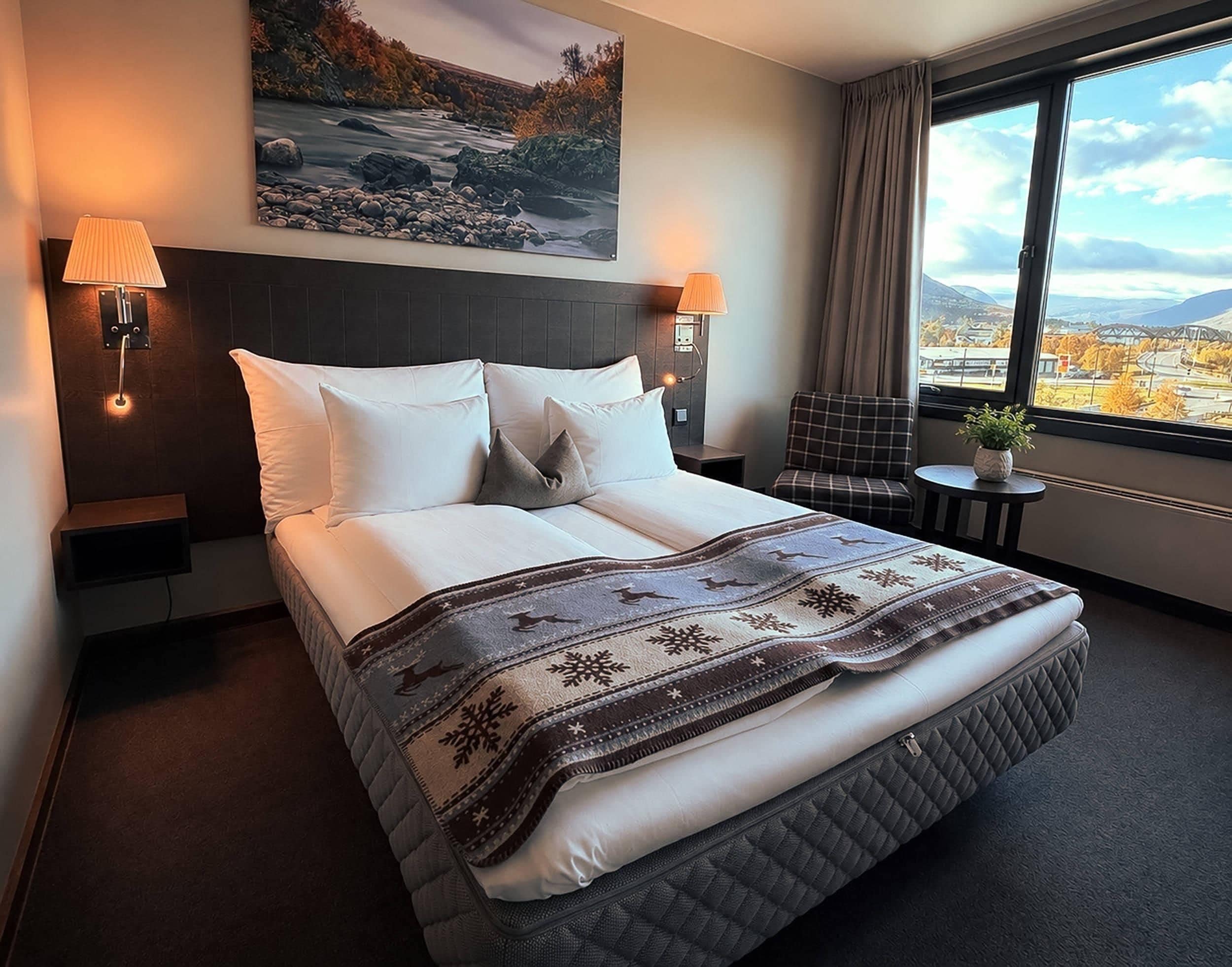 A hotel room with a bed, two bedside lamps, a chair, a small table, a window with a view, and a painting on the wall.