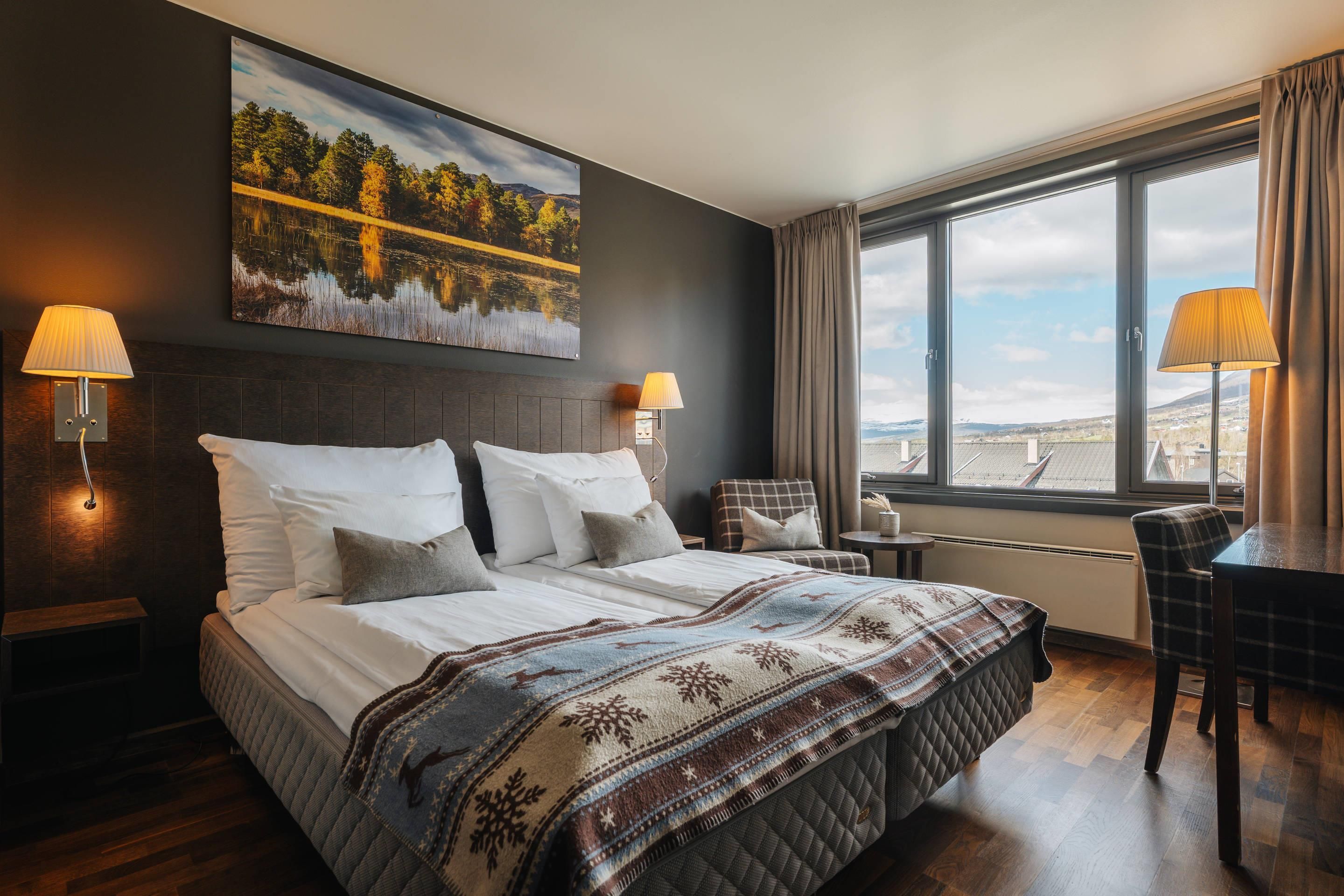 A hotel room with a large bed, two bedside lamps, a view of the landscape through the window, and a painting on the wall.