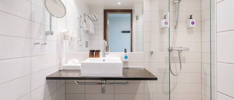Un baño moderno con pared de azulejos blancos, encimera negra, espejo, lavamanos, cabezal de ducha manual y mampara de cristal.