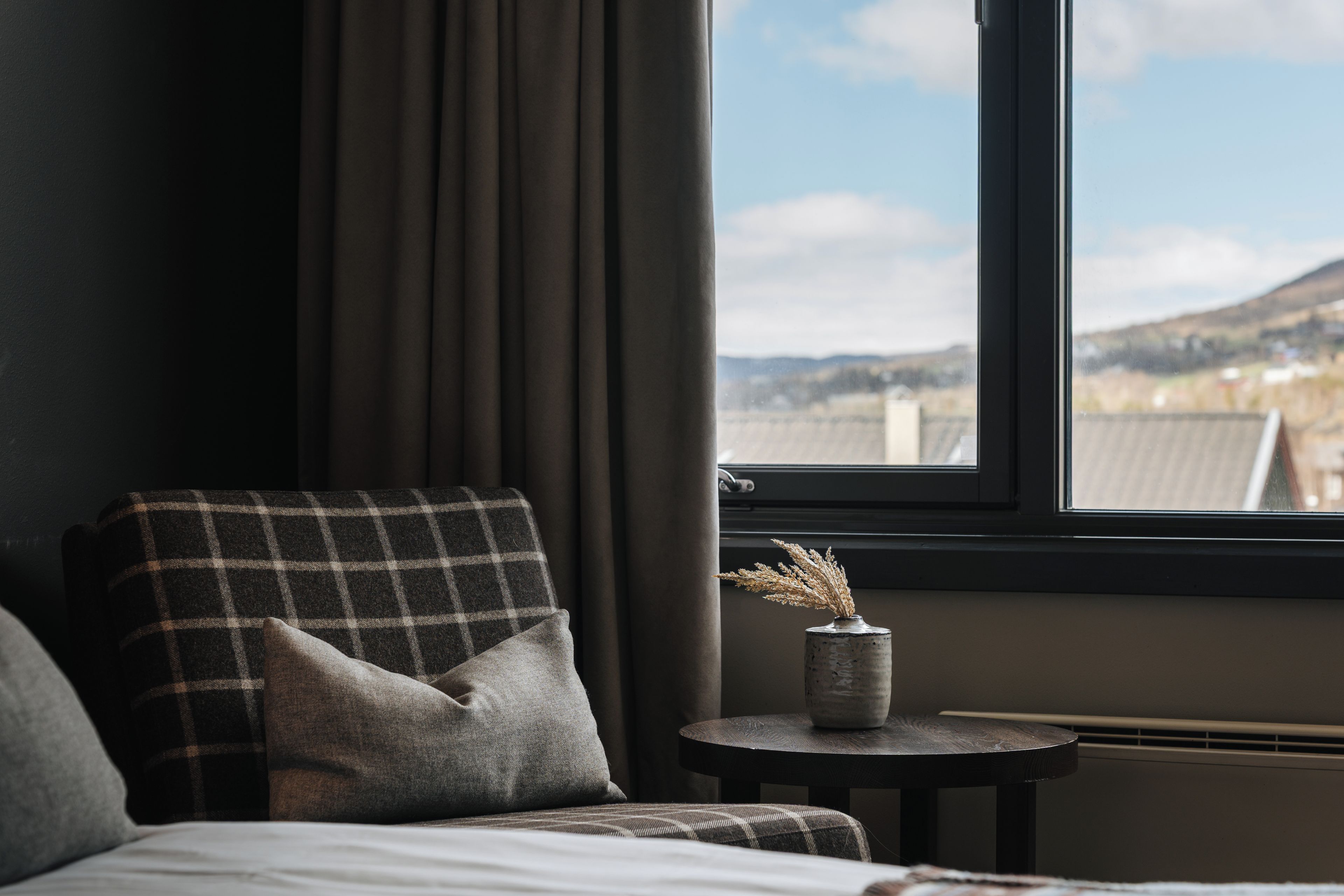 A room with a bed, a chair, a small table with a vase, and a window with a view of the outdoors.