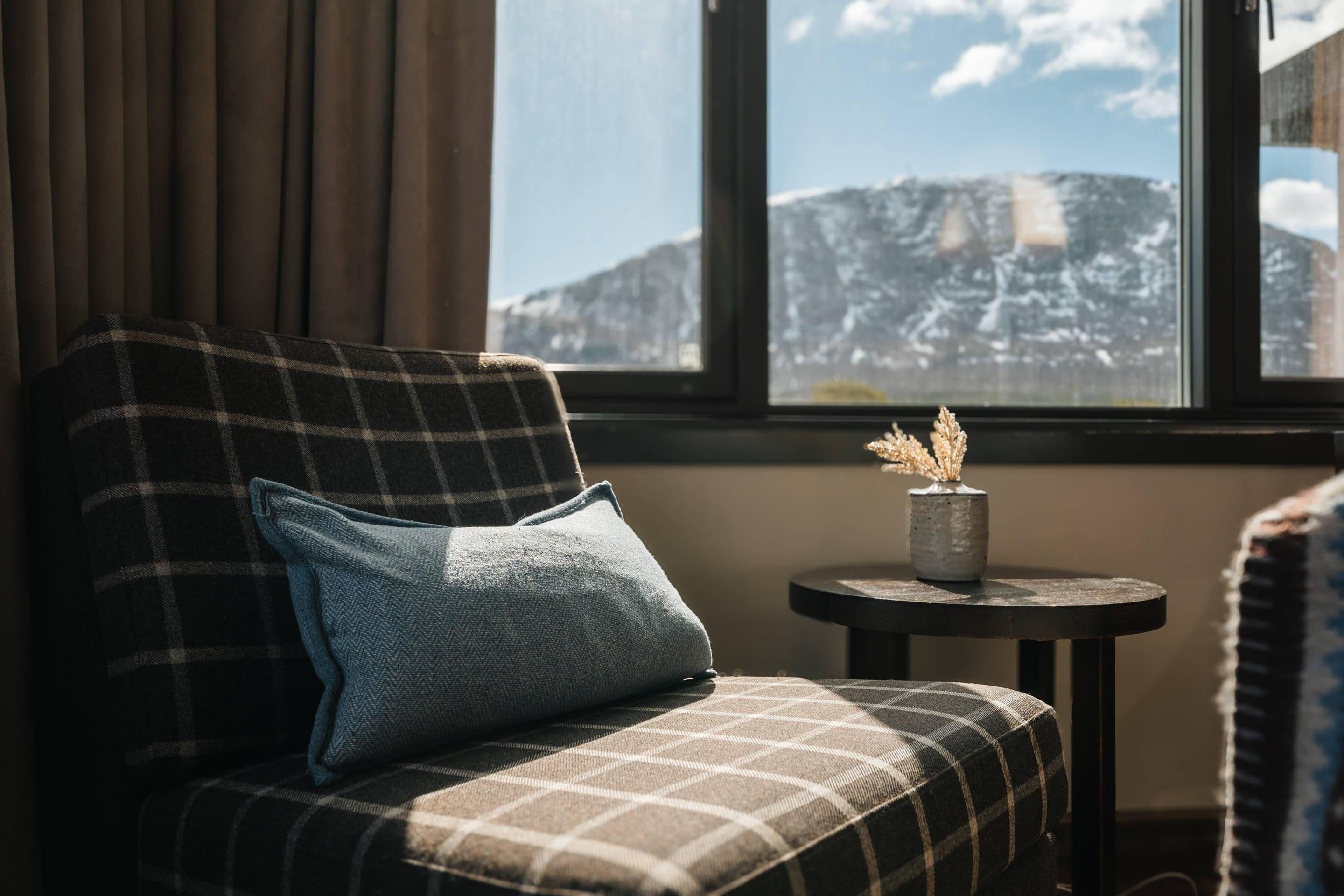 A plaid sofa with a cushion, a small round table with a vase, and a window with a mountain view.