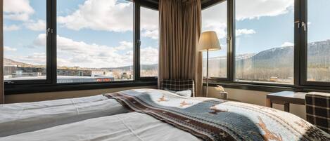 Habitación de hotel con una cama grande, un escritorio y una lámpara. Varias ventanas ofrecen vistas a las montañas y al paisaje urbano.