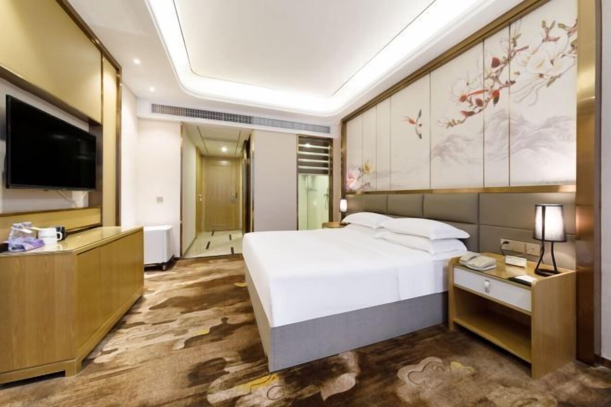 A modern hotel room with a large bed, a TV mounted on the wall, a desk with a lamp, and a decorative wall panel.