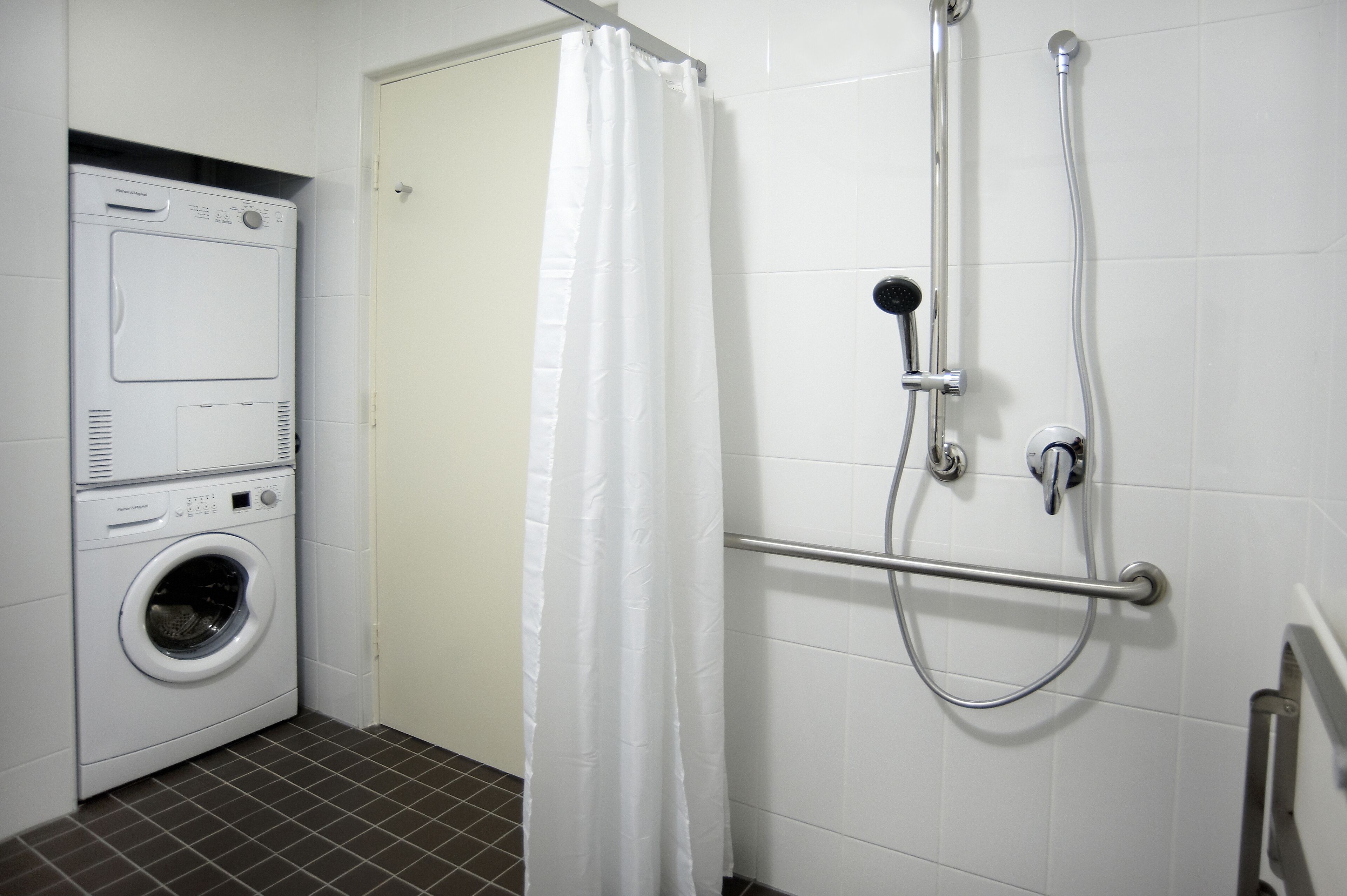 A laundry area with a washing machine and a shower with a grab bar.
