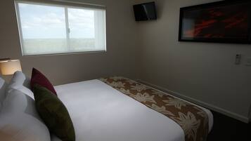 Habitación de hotel con cama, almohadas, ventana, televisor montado en la pared y un cuadro enmarcado.