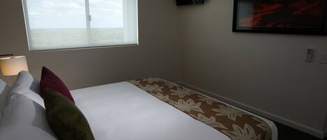 Habitación de hotel con cama, almohadas, ventana, televisor montado en la pared y un cuadro enmarcado.