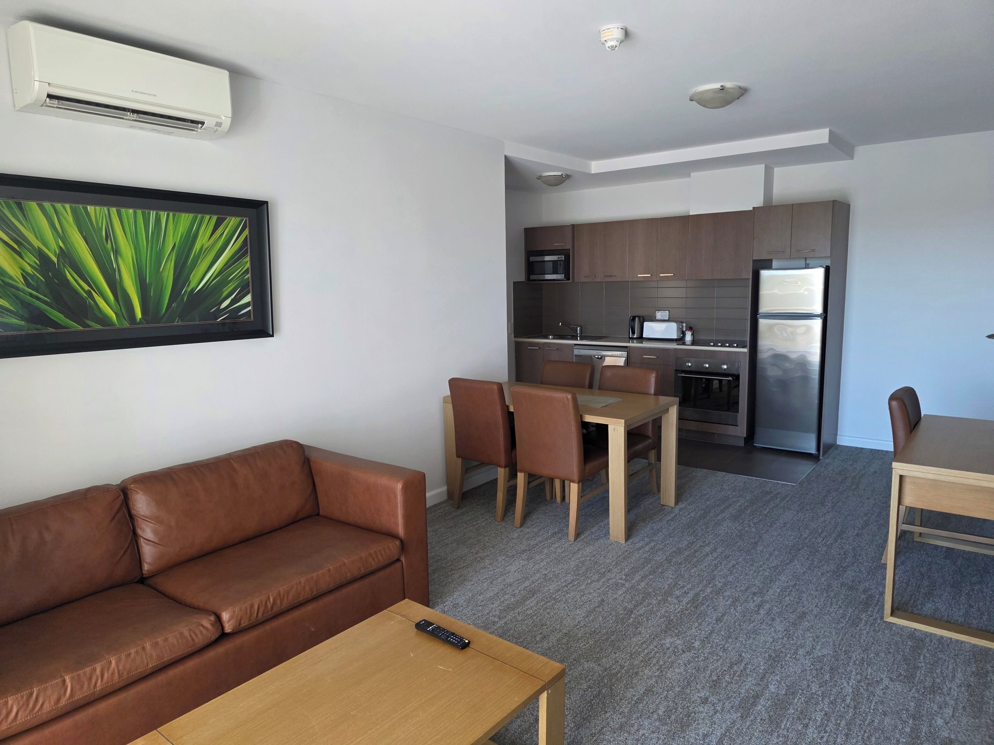 Una sala de estar moderna con un sofá de cuero marrón, una mesa de centro de madera y un comedor con mesa y sillas. Se ve una cocina al fondo con electrodomésticos modernos.