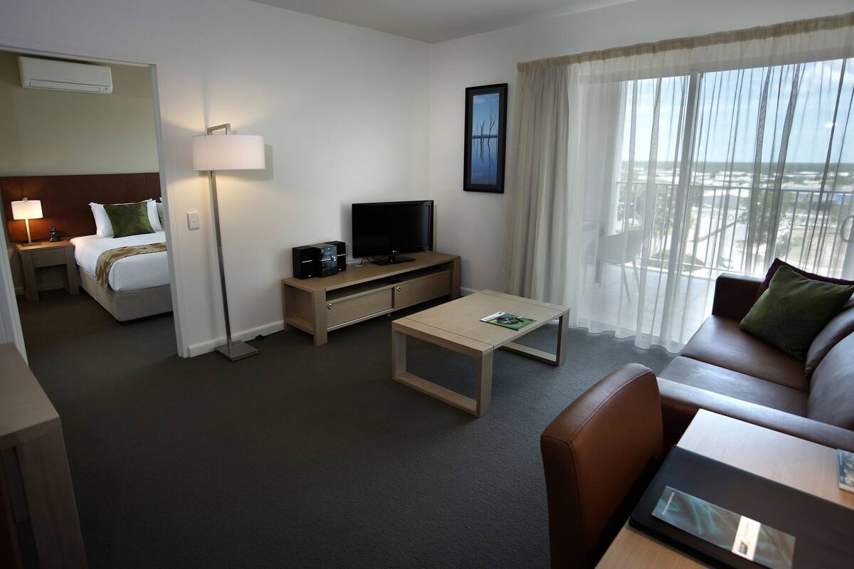 Habitación de hotel con cama, televisor, sofá, mesa de centro y ventana con cortinas.