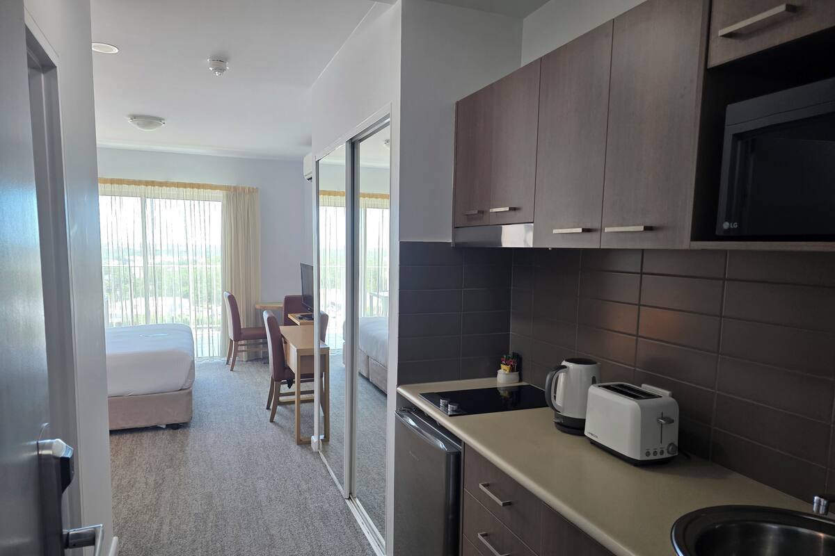 Habitación de hotel con kitchenette, zona de comedor con mesa y sillas, cama y un amplio ventanal con cortinas.