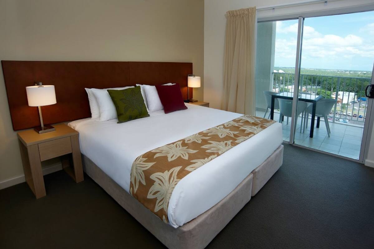 Habitación de hotel con una cama grande, dos mesitas de noche con lámparas, un balcón con mesa y sillas, y vistas a la ciudad.