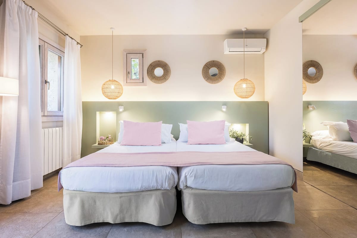Una cama doble con dos almohadas, una mesita de noche con lámpara, una ventana con cortinas y un aire acondicionado instalado en la pared.