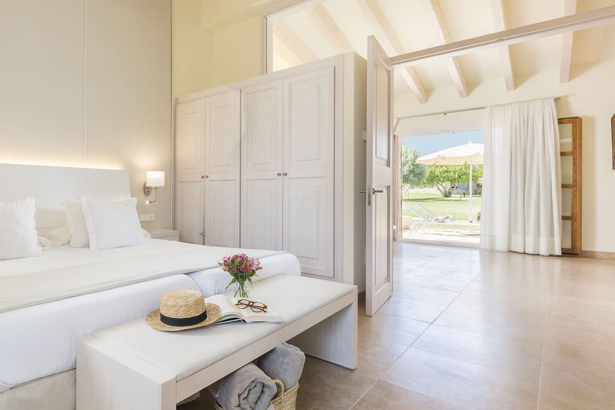 Un dormitorio con una cama grande, una mesita de noche con un sombrero y gafas, un armario y una puerta abierta que da a un balcón.