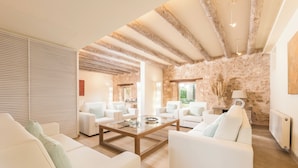 Un salón amplio con sofás blancos, una mesa de centro de cristal, vigas de madera a la vista y una pared de piedra.