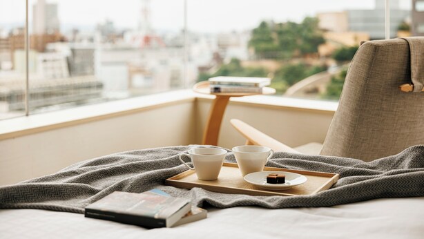 A cozy room with a city view, a bed with a blanket, a tray with cups and a book, and a chair. - Fraser Residence Nankai Osaka (Osaka)
