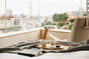 Une pièce confortable avec vue sur la ville, un lit avec une couverture, un plateau avec des tasses et un livre, et une chaise.