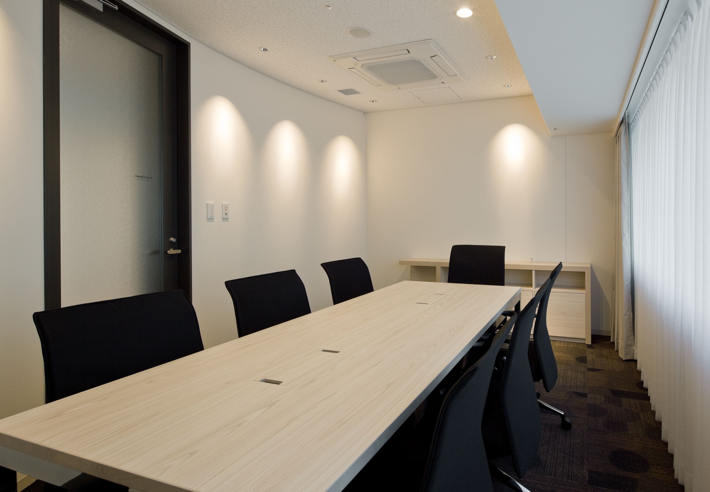 A conference room with a long table, black chairs, a white desk, and a door with frosted glass.