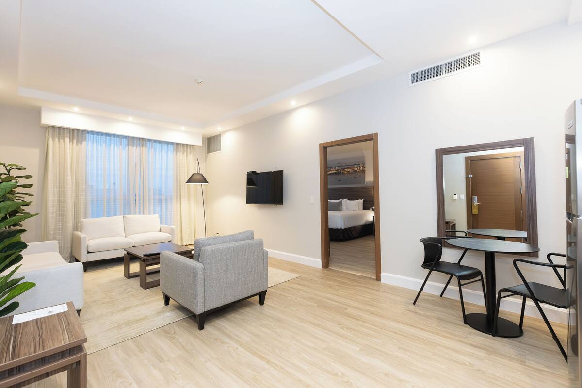 Een moderne hotelkamer met een zithoek, een eettafel en een slaapkamer, zichtbaar door een geopende deur.