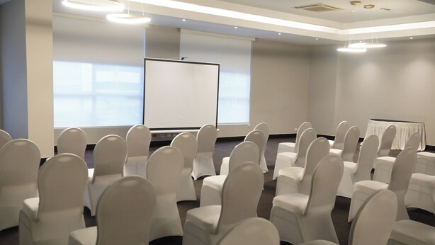 Een vergaderzaal met rijen witte stoelen, een scherm voor presentaties en een groot raam.
