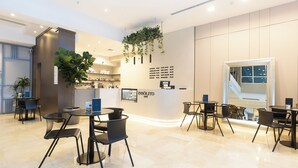 Een modern café met een strak, minimalistisch design, met een balie met een menukaart, verschillende tafels en stoelen, en een grote plant die van het plafond hangt.