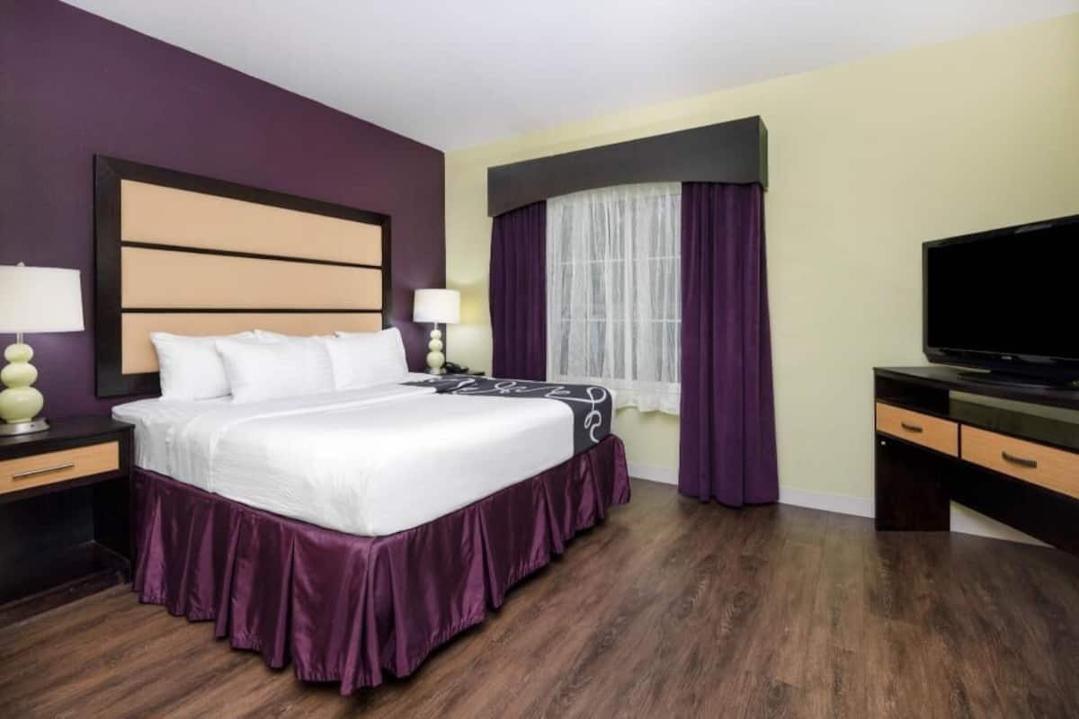 Ein Hotelzimmer mit einem großen Bett, einem Schreibtisch mit Fernseher, einem Nachttisch und einem Fenster mit violetten Vorhängen.