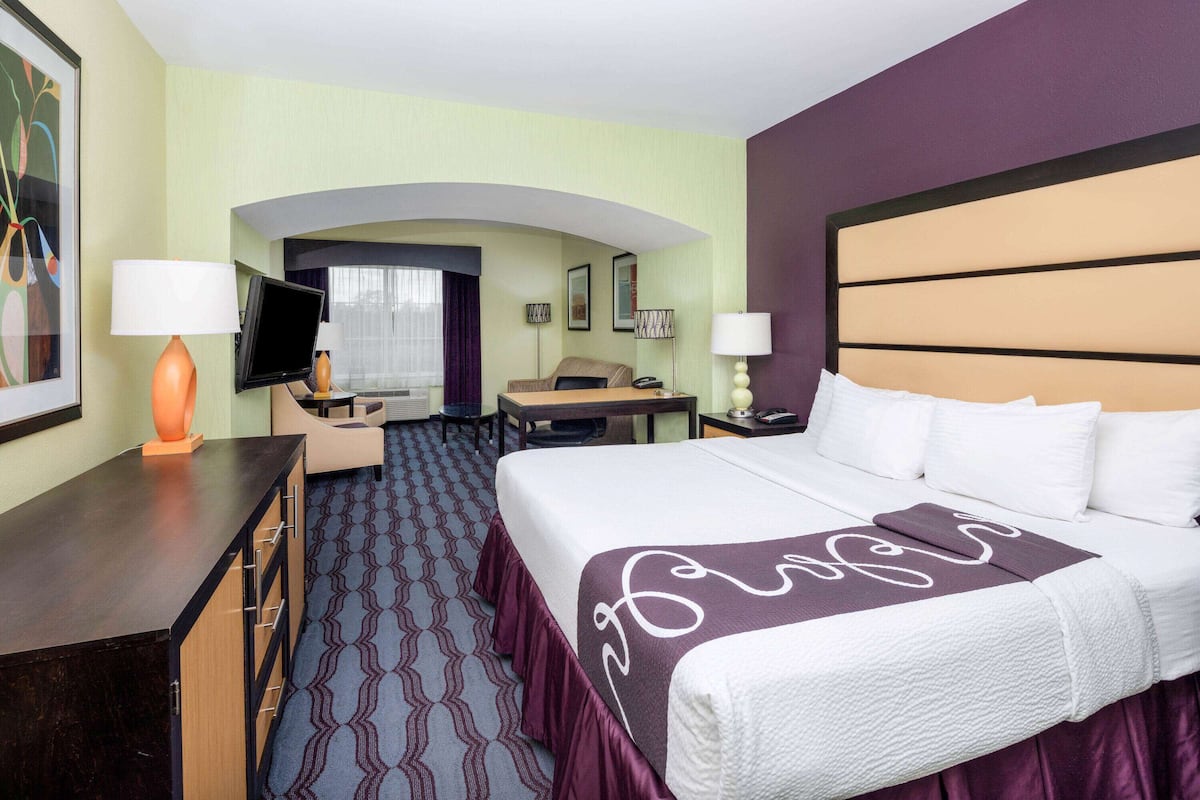 Ein Hotelzimmer mit einem großen Bett, einem Schreibtisch mit Lampe, einem Fernseher und einer Sitzecke mit Couch und Sesseln.
