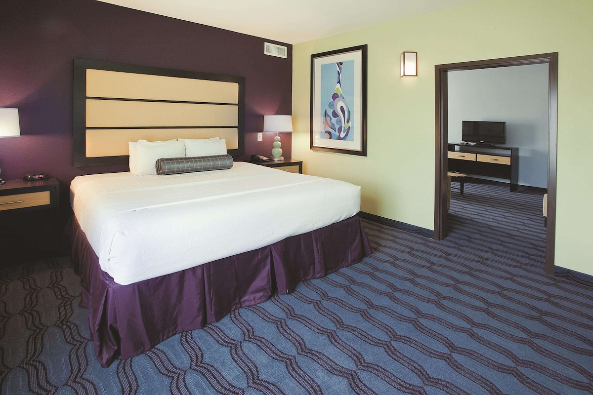 Ein Hotelzimmer mit einem großen Bett, einem Schreibtisch mit Fernseher und einer Tür zu einem weiteren Raum.