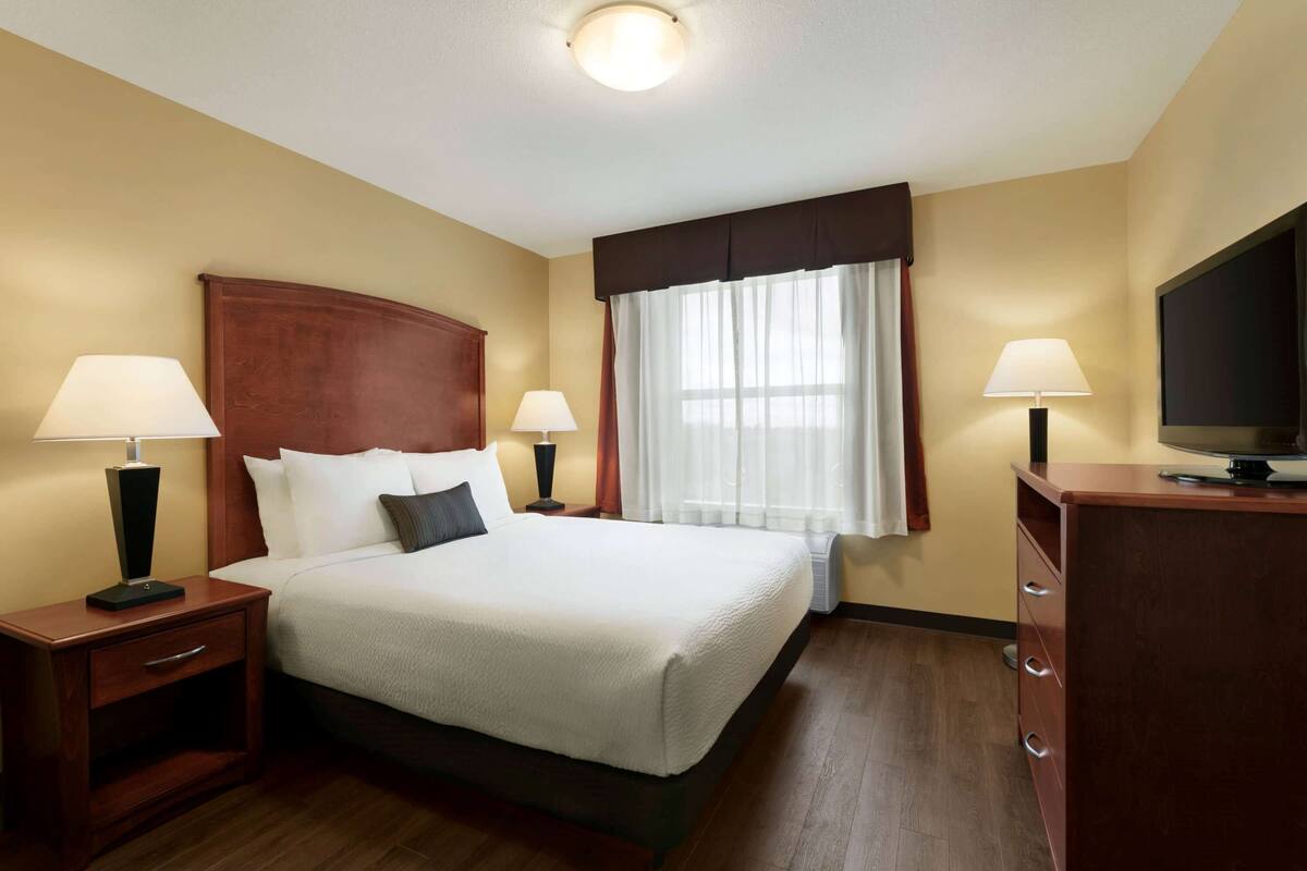 Una habitación de hotel con una cama grande, dos mesitas de noche con lámparas, un televisor en un aparador y una ventana con cortinas.