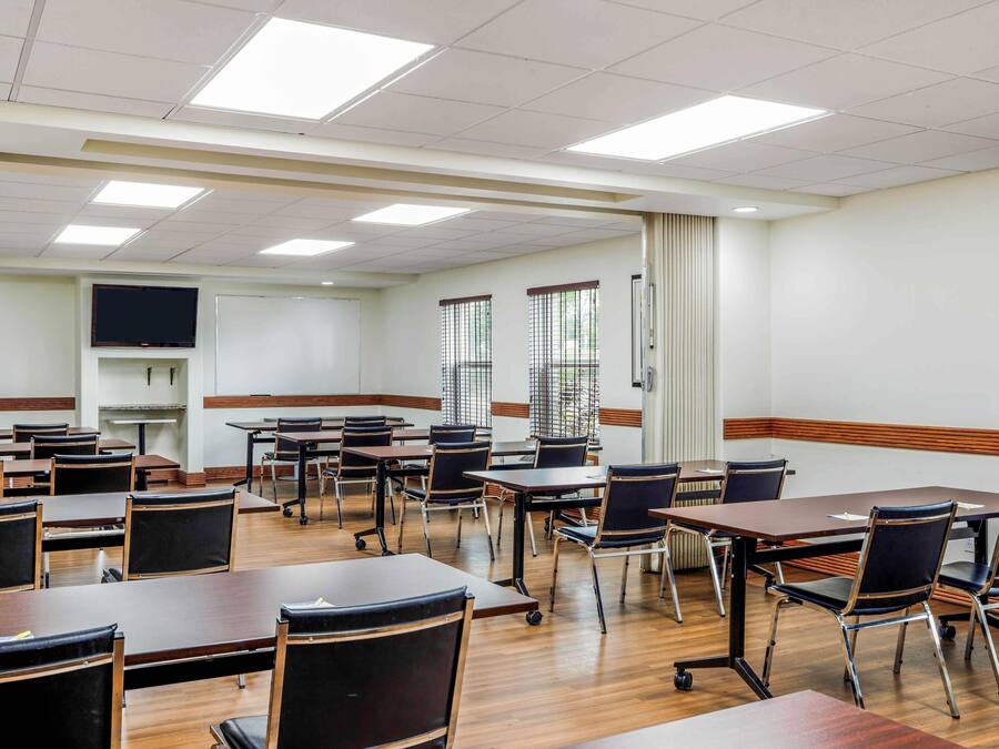 Un aula con mesas y sillas de madera, una pizarra y ventanas con persianas.
