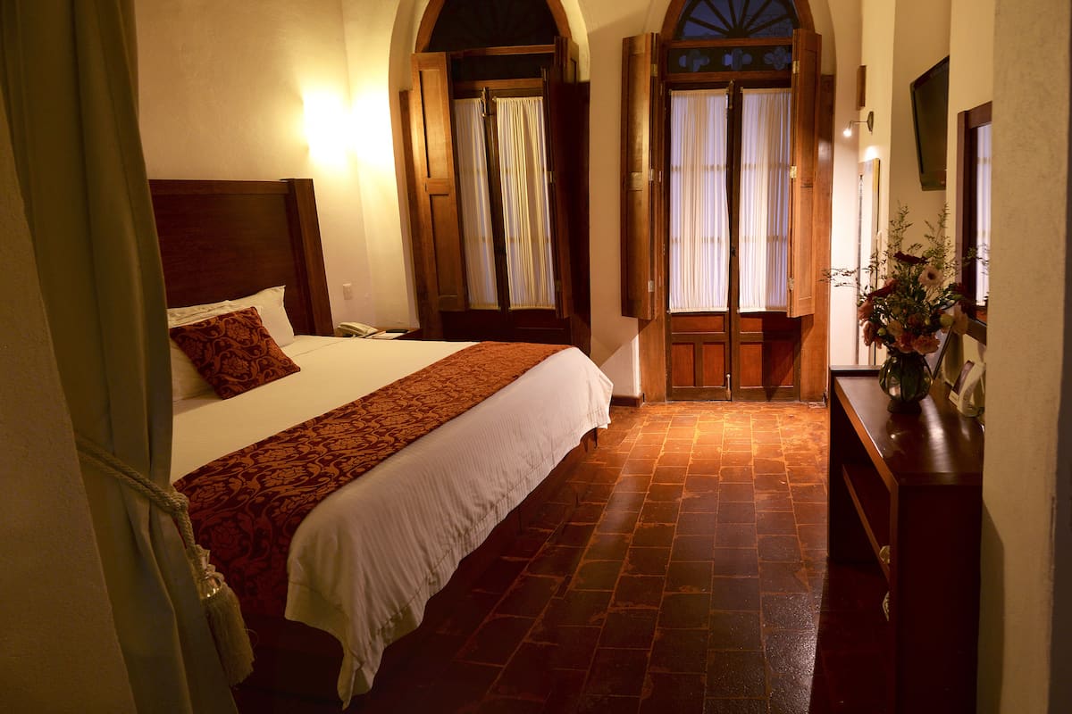 A hotel room with a large bed, a wooden headboard, a stone-tiled floor, a wooden door with a window, and a vase with flowers on a table.