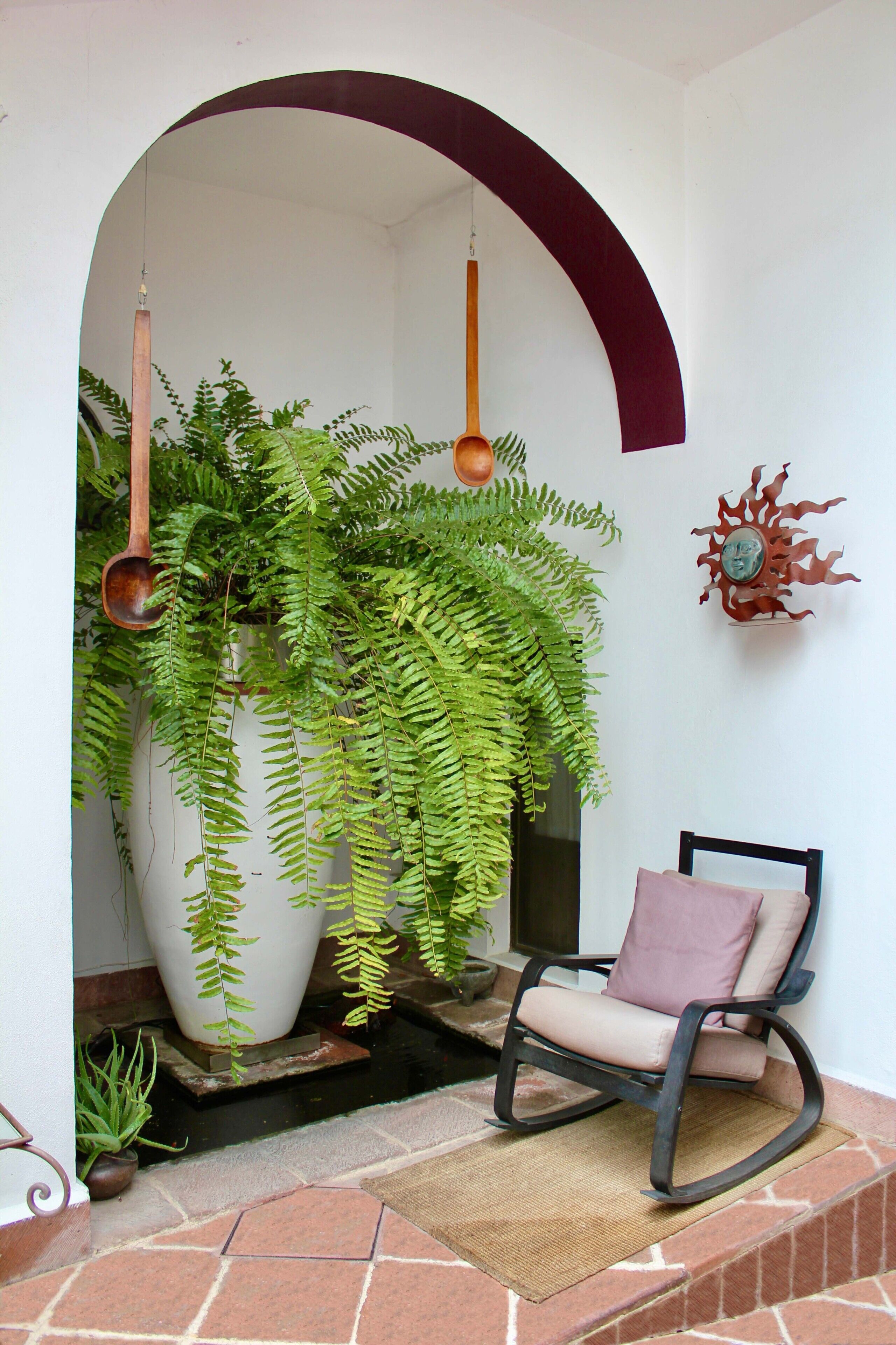 A patio area with a white wall, a large potted plant, a rocking chair, and a decorative metal piece.