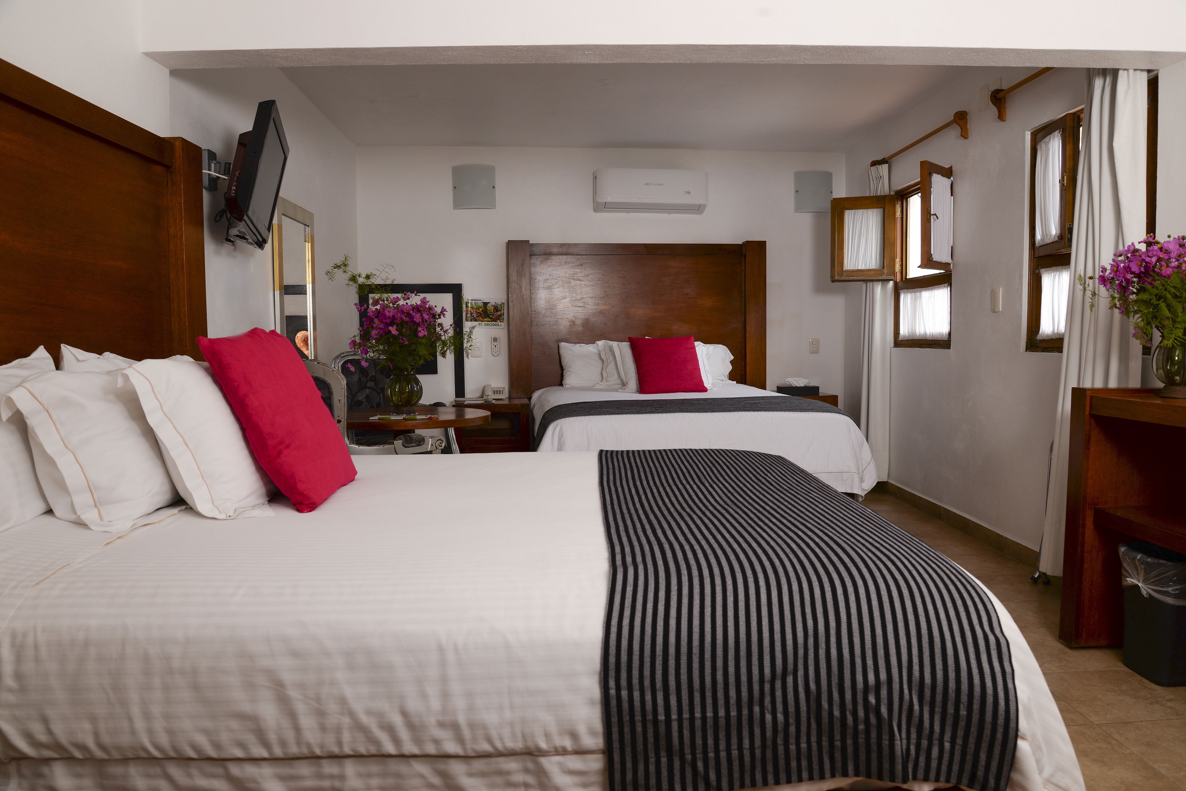 Ein Hotelzimmer mit zwei Betten, einem Fernseher, einer Klimaanlage und einem Schreibtisch mit Blumen.