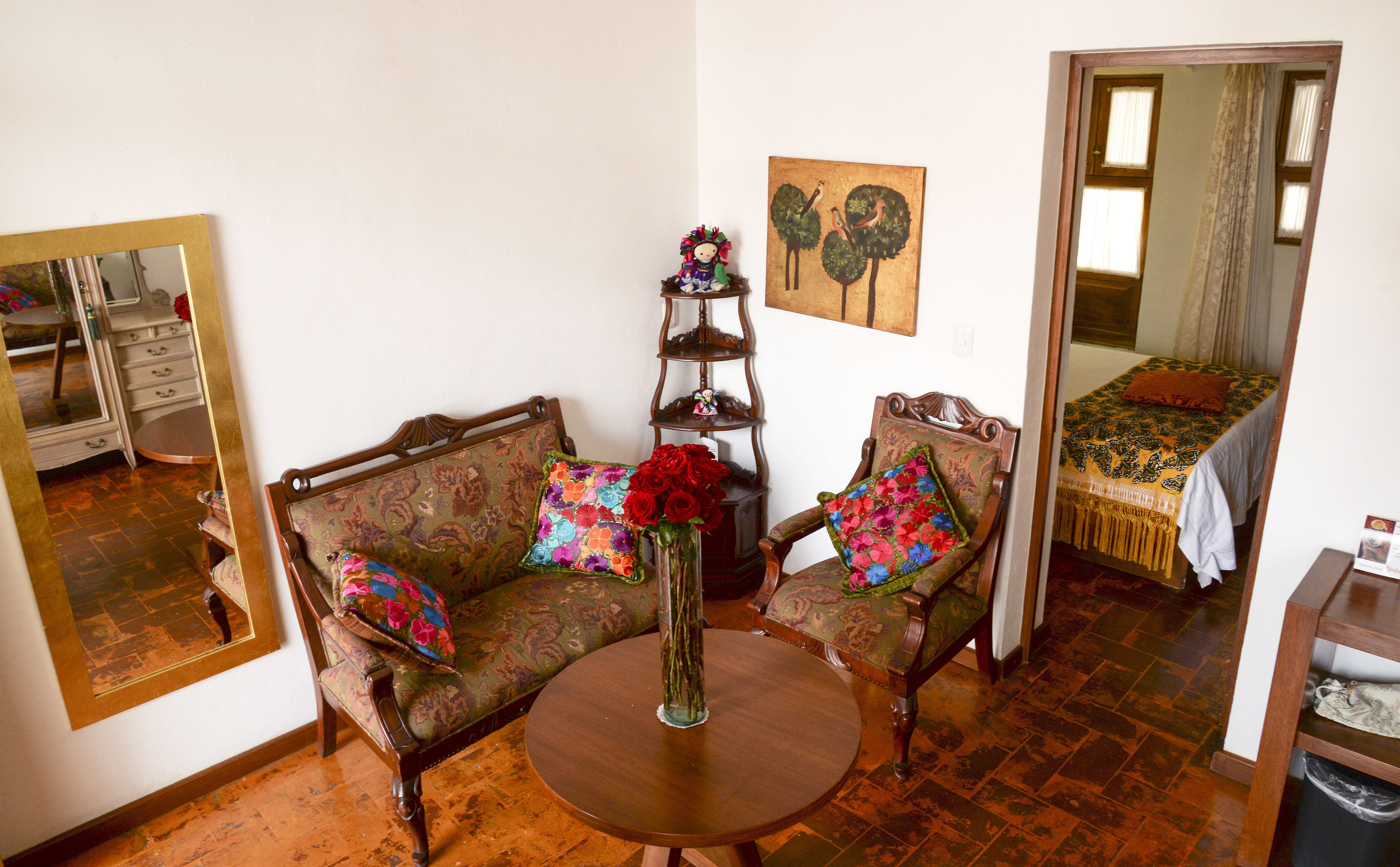 A room with a sofa, a chair, a round table, a mirror, a bed with a patterned bedspread, and a small shelf with decorative items.