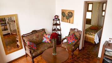 A room with a sofa, a chair, a round table, a mirror, a bed with a patterned bedspread, and a small shelf with decorative items.