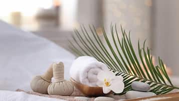 Un ambiente de spa con una camilla de masaje, una toalla blanca enrollada, un cuenco de madera con una flor blanca y una hoja de palma.