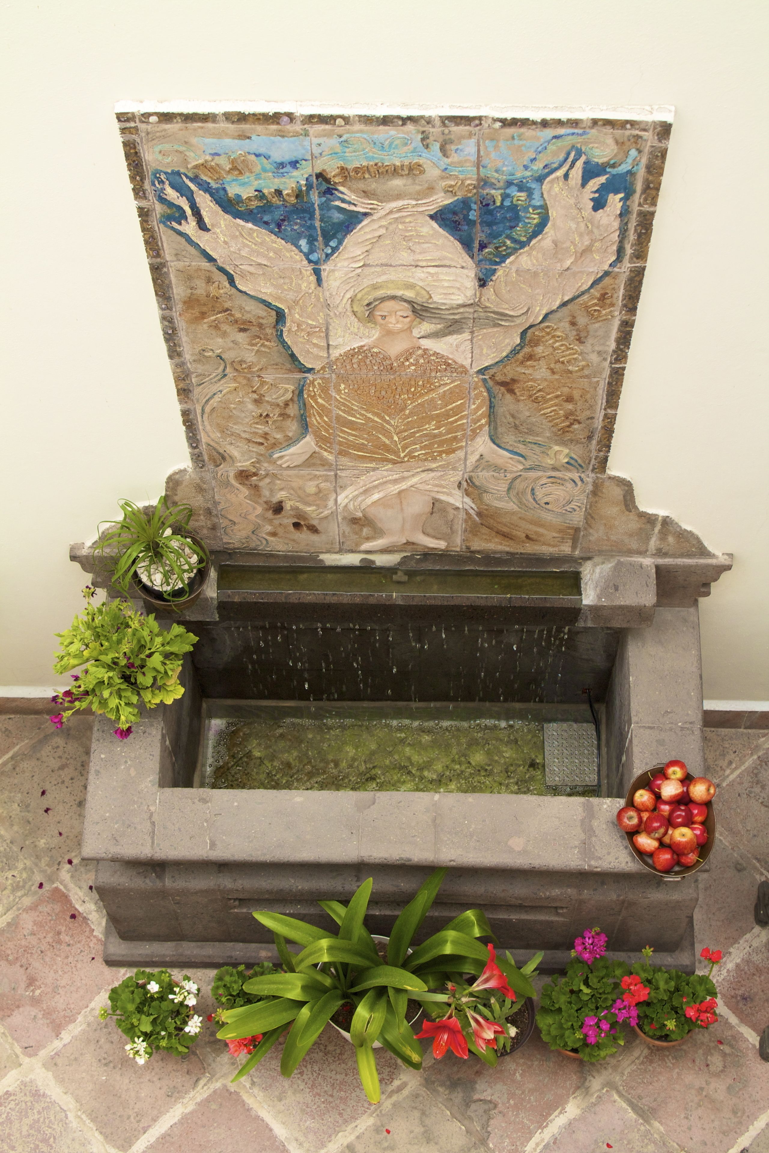 Una fuente de piedra con un ángel en mosaico, rodeada de plantas en macetas y manzanas.