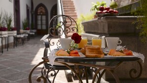 Una mesa de desayuno con tostadas, fruta y jugo en un patio con suelo de piedra y mobiliario exterior.