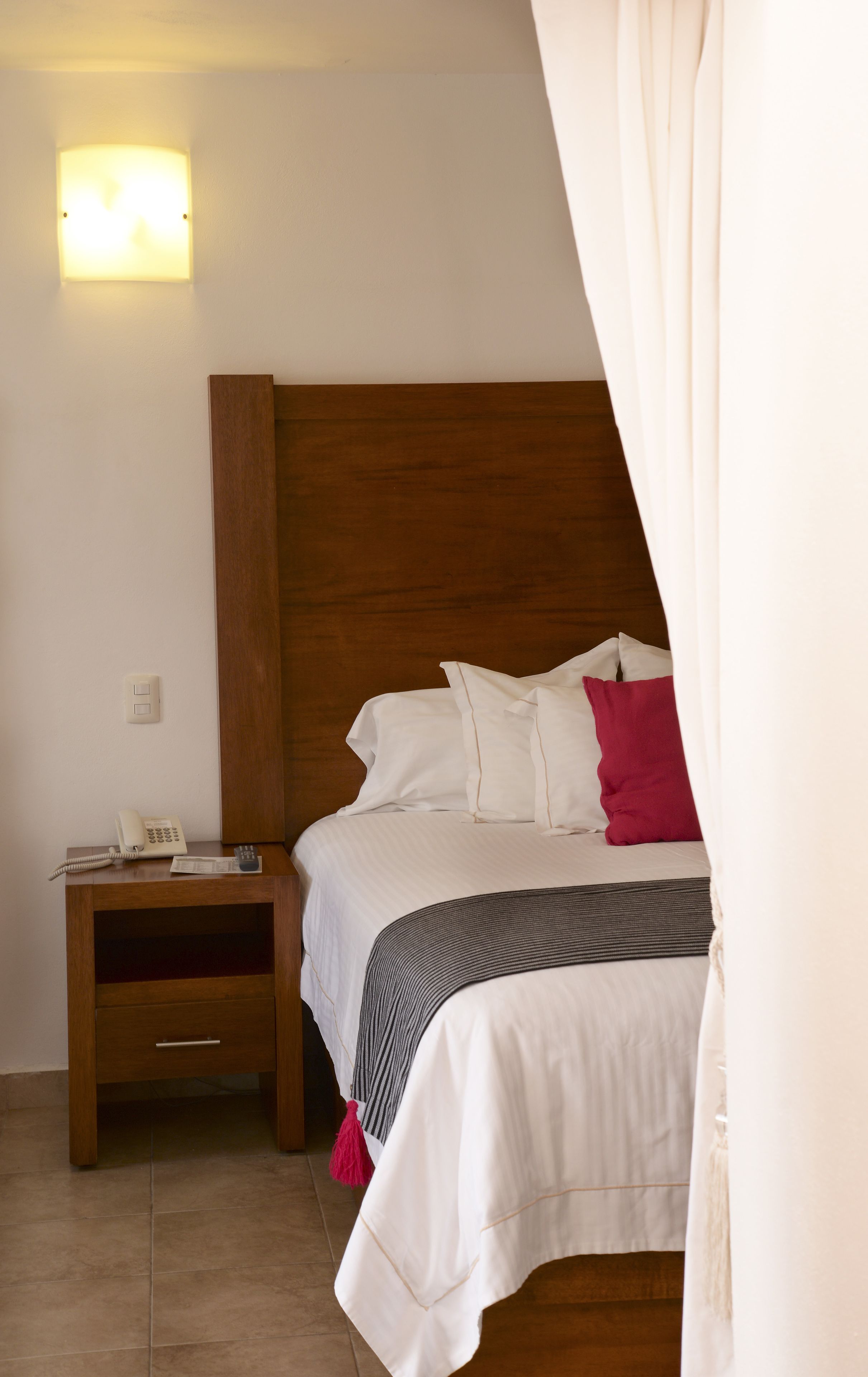 Ein Schlafzimmer mit einem hölzernen Kopfteil, einem Nachttisch mit Telefon sowie einem Bett mit weißer Bettwäsche und einem roten Kopfkissen.