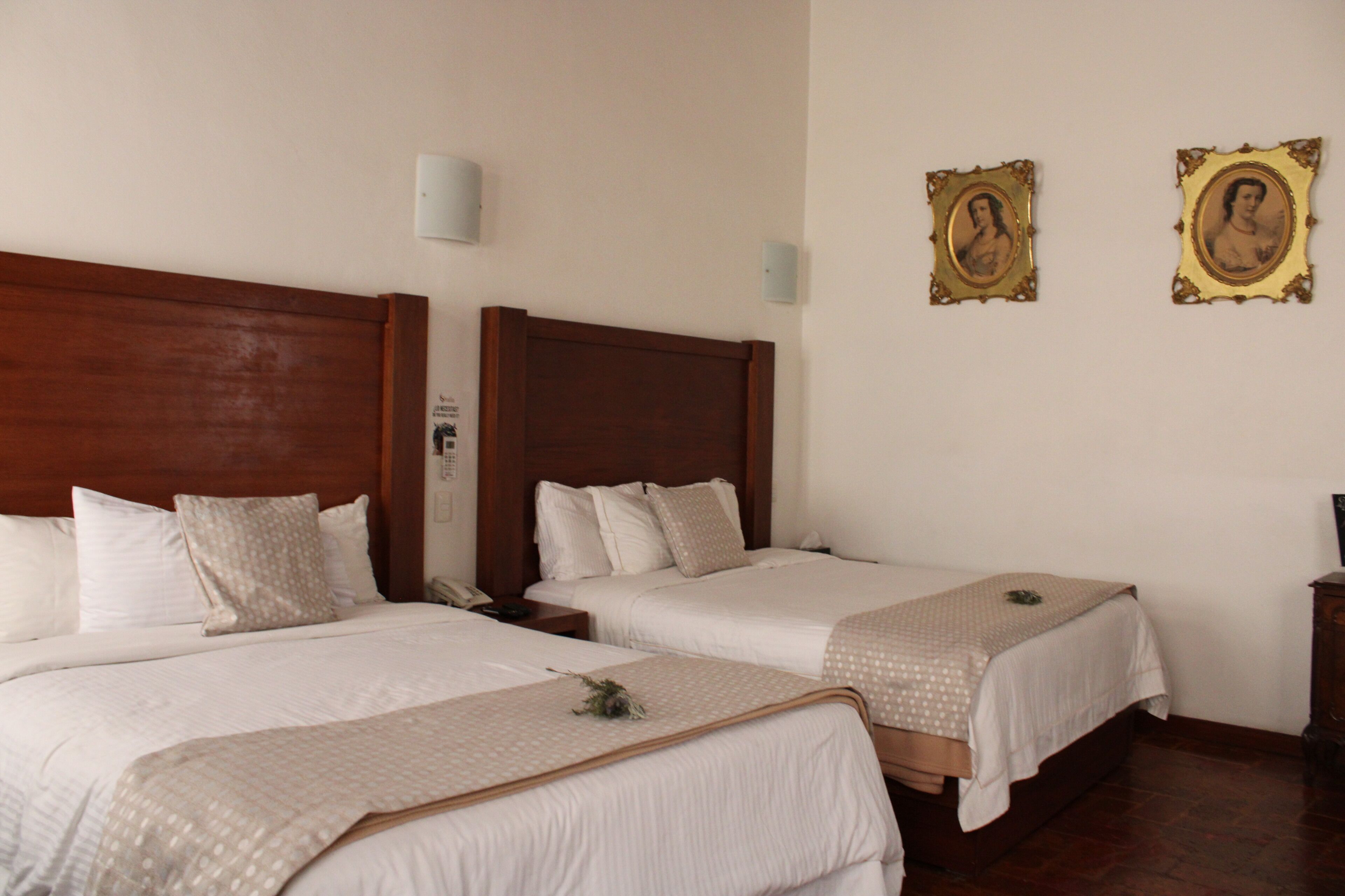 Una habitación de hotel con dos camas individuales, cabeceros de madera, ropa de cama blanca y dos retratos enmarcados en la pared.