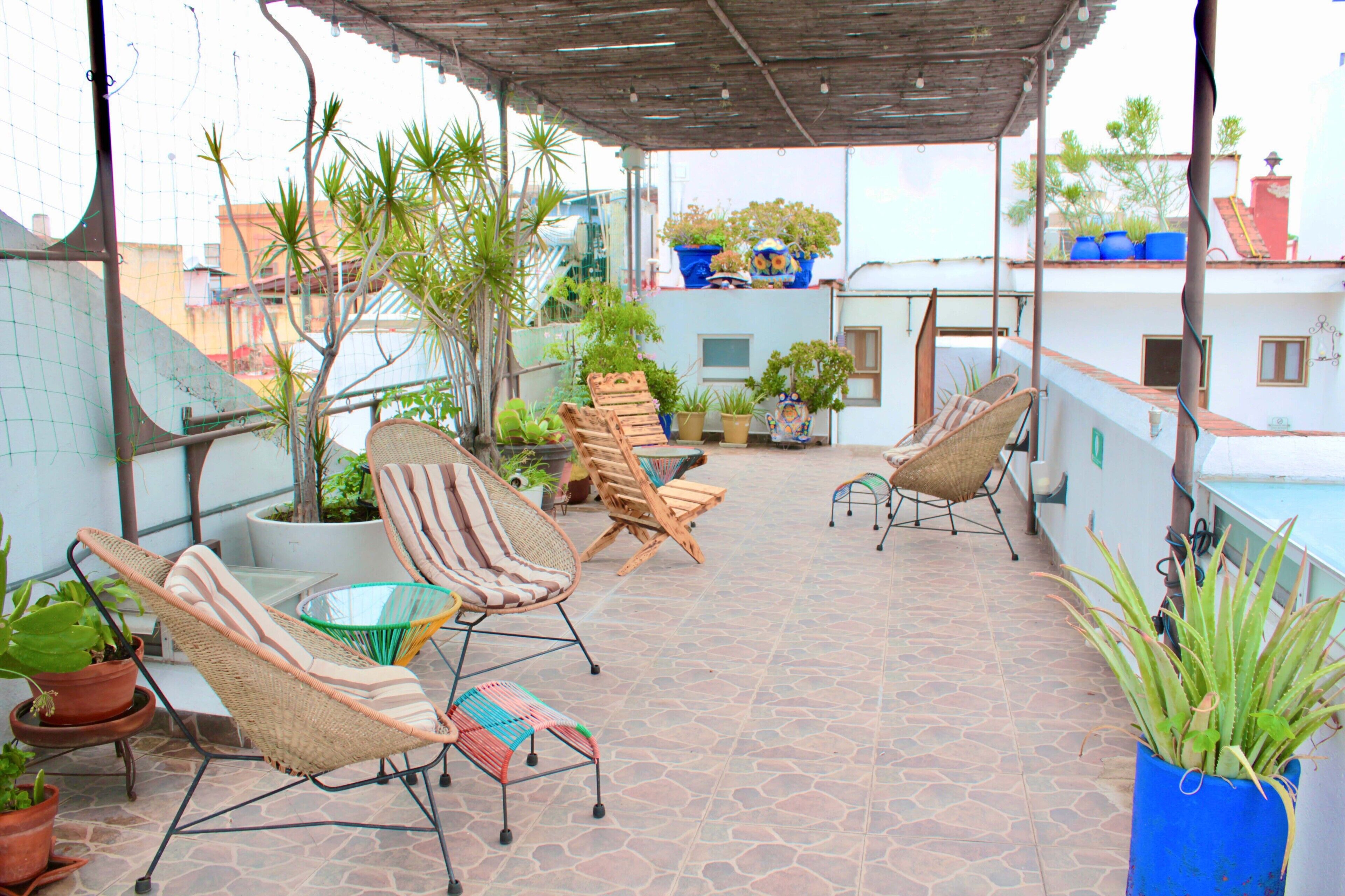 Un espace extérieur aménagé avec des meubles de jardin en osier, comprenant des chaises et des tables, le tout protégé par un auvent et agrémenté de plantes en pot et d’un sol carrelé.