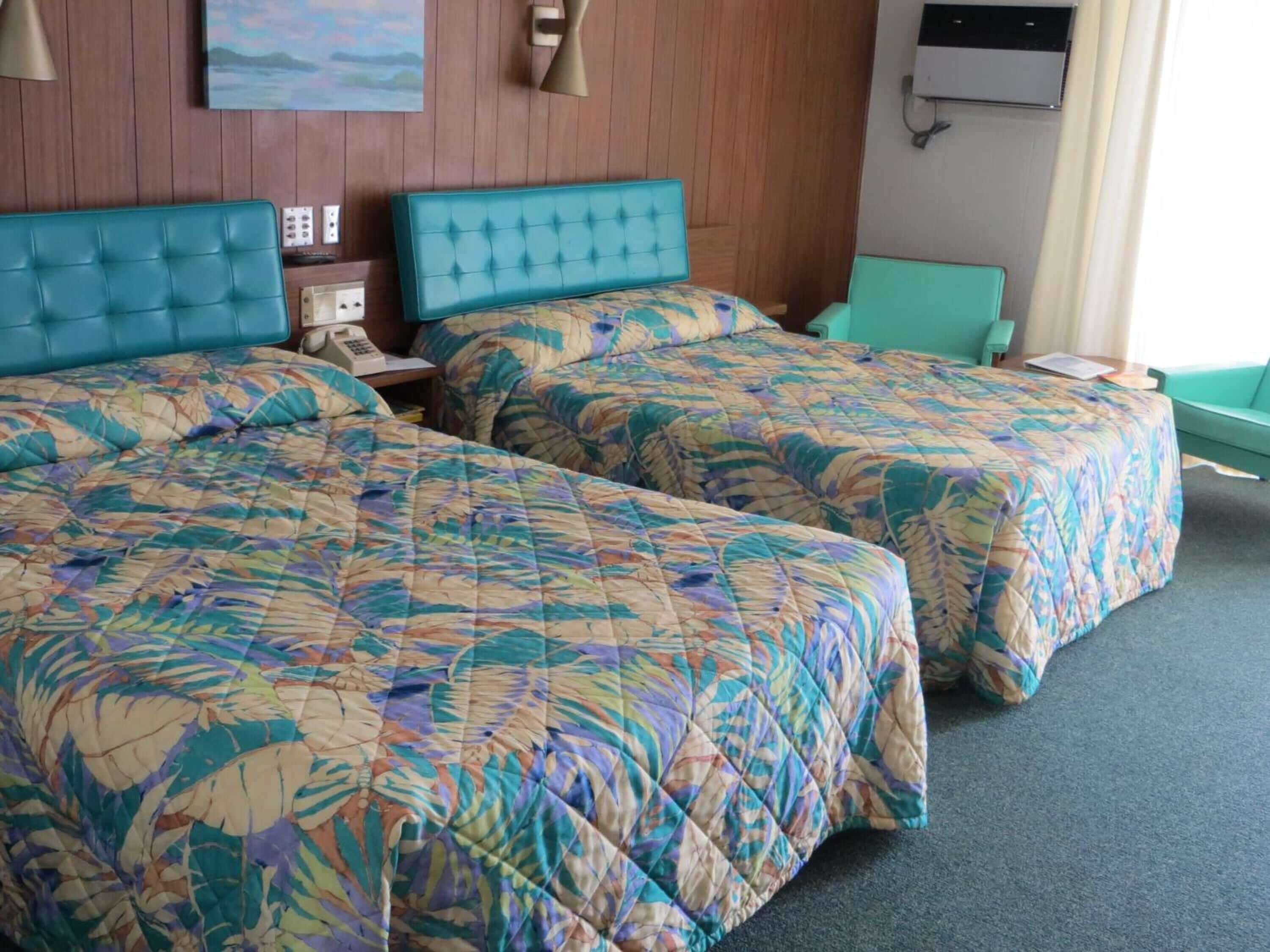 A hotel room with two beds, each with a floral-patterned bedspread, a wooden headboard, a bedside table with a lamp, and a painting on the wall.