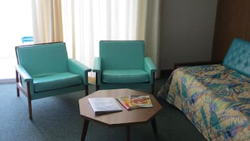 Quarto de hotel com duas poltronas azuis, uma mesa de centro de madeira com revistas e uma cama com colcha estampada de flores.