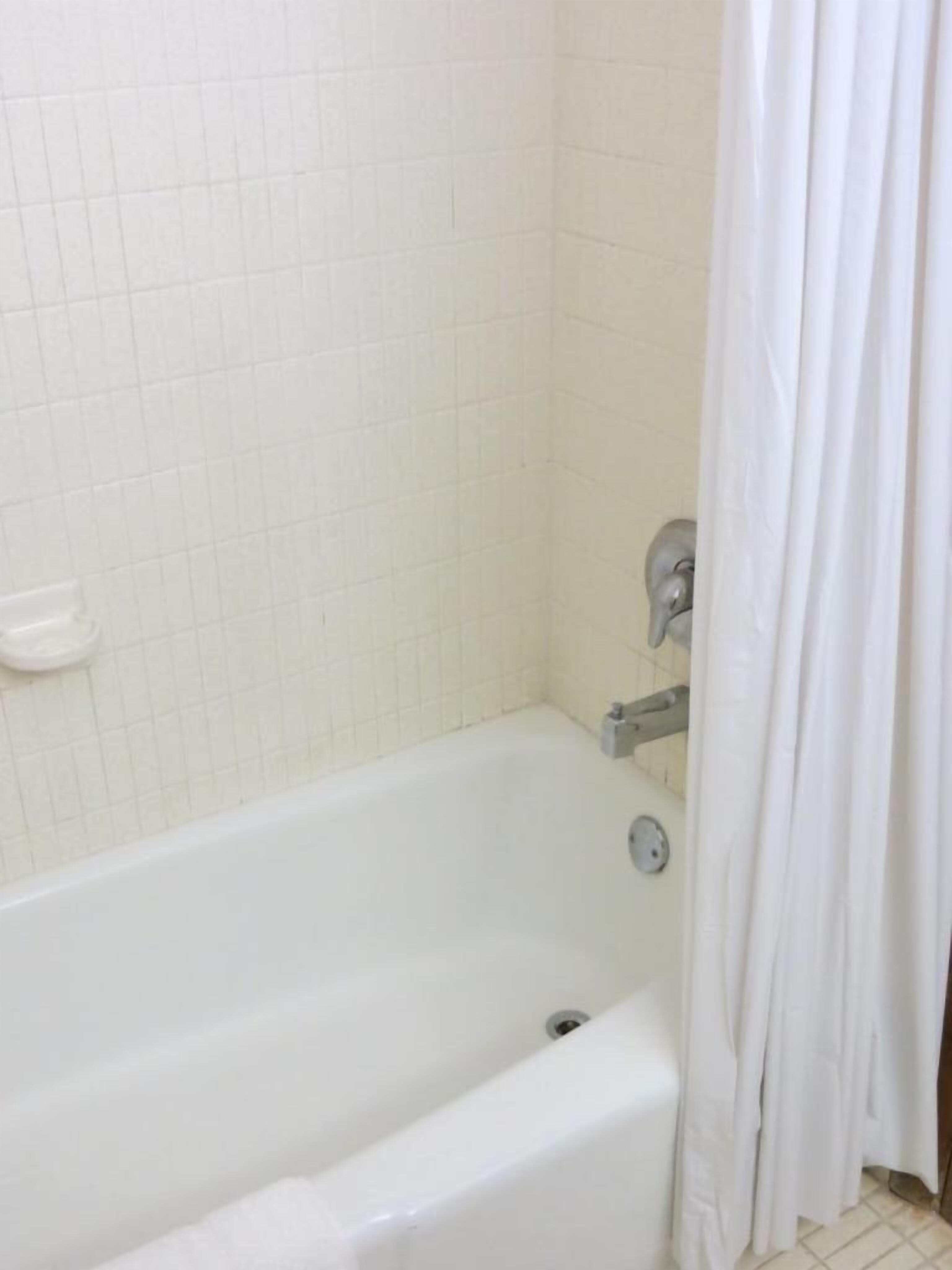 Eine weiße Standardbadewanne mit Duschvorhang, einer kleinen weißen Ablage und einer gefliesten Wand.