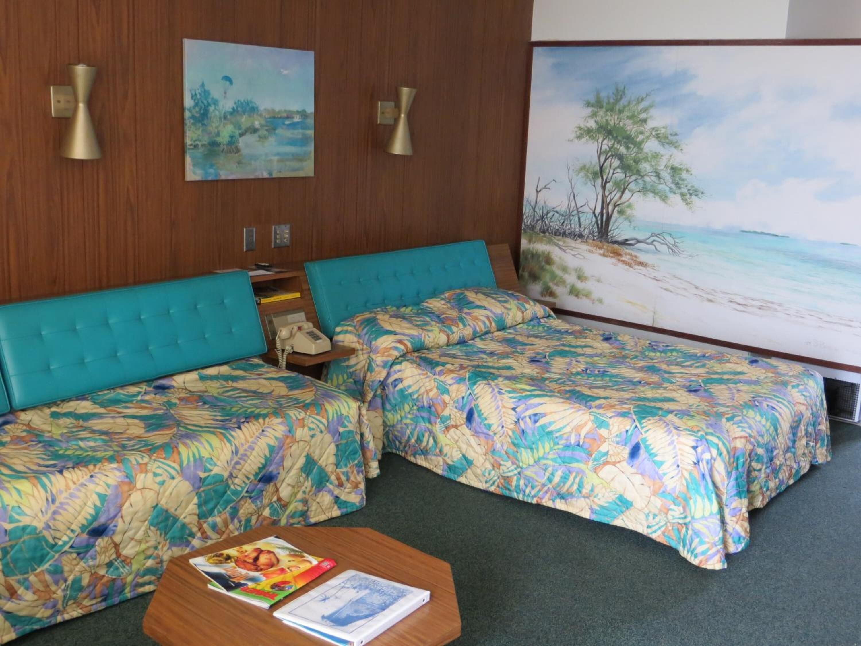 A hotel room with two beds, a small table, and a painting of a beach scene.