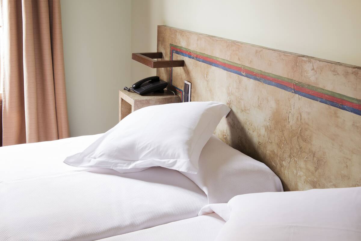Una cama bien hecha con sábanas y almohadones blancos, una mesita de noche de madera con un teléfono, y una pared con diseño rústico y sencillo.