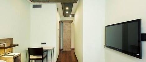 A hotel room with a bed, a desk with a chair, a television mounted on the wall, and a hallway leading to another room.