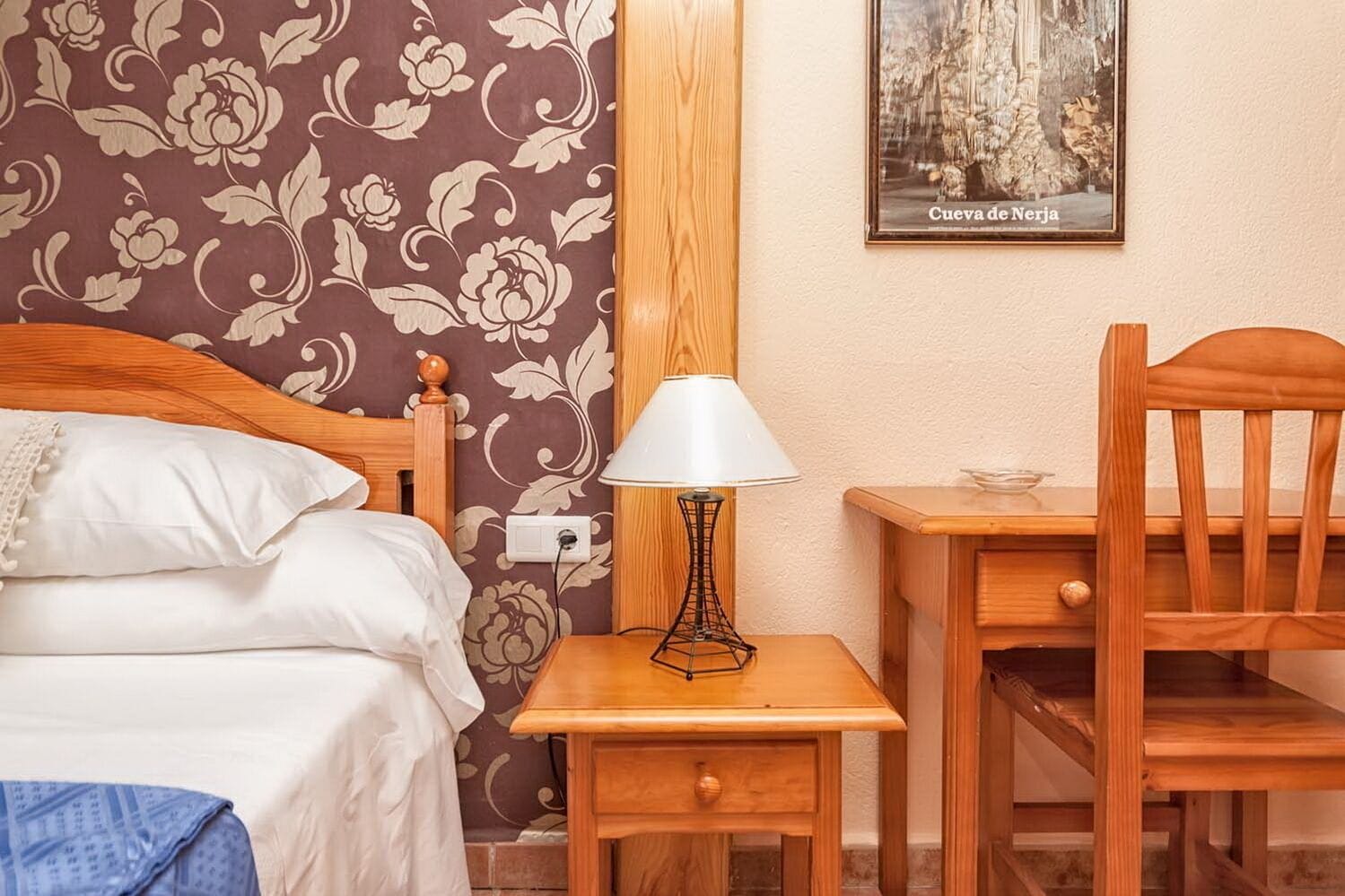 A bedroom with a bed, a nightstand, a table, and a chair. There is a lamp on the nightstand, and a framed picture on the wall.
